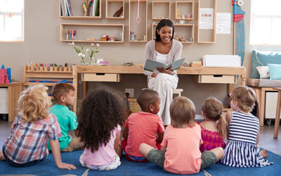 Children sitting on the floor, listening attentively as a teacher reads them a story at Little Lights.